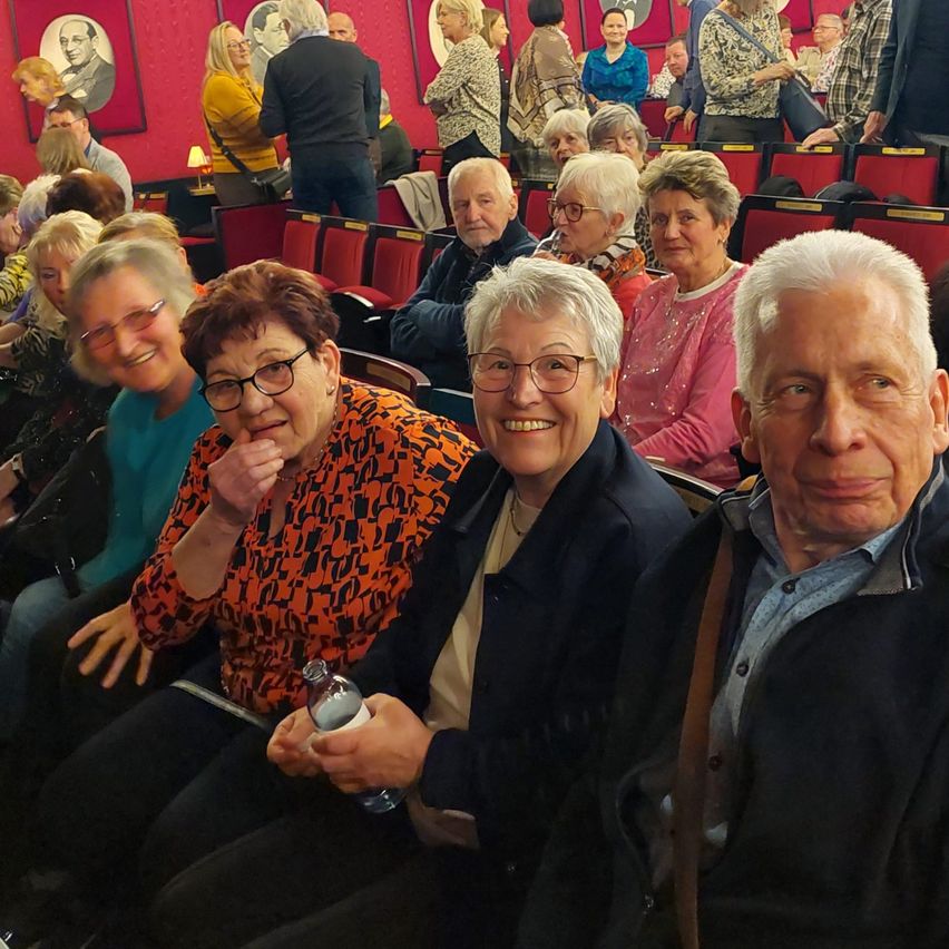 Eine Gruppe älterer Erwachsener sitzt in einem Theater. Sie alle lächeln und genießen wahrscheinlich eine Aufführung. Ein Mann hält eine Flasche.