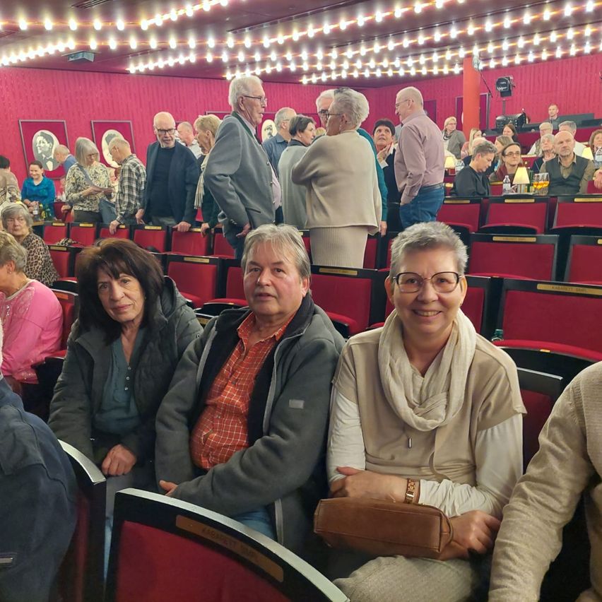 Eine Gruppe von Menschen sitzt in einem Theater, einige mit Brille und lächelnd. Hinter ihnen steht ein Publikum im Gang. Das Theater hat rote Sitze und eine rote Wand mit gerahmten Bildern.