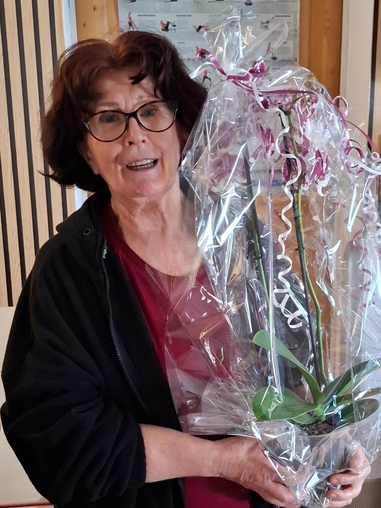 An older woman with glasses smiles and holds a wrapped plant. The plant is in a pot with green leaves. She is standing in front of a wooden wall.