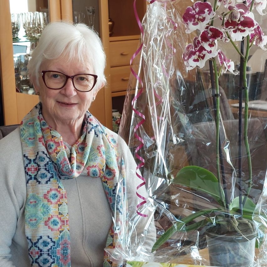 Eine ältere Frau mit weißen Haaren und einer Brille sitzt neben einer in Plastik eingewickelten Orchideenpflanze mit rosa und weißen Blüten.