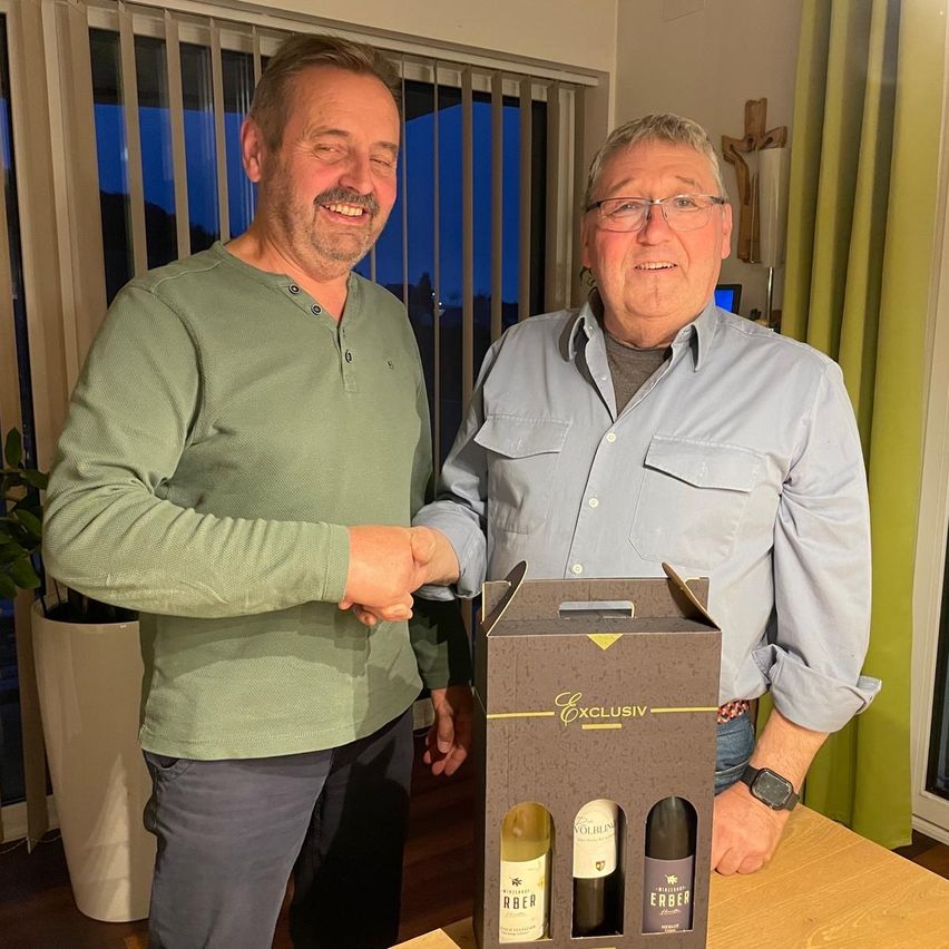 Zwei Männer stehen in einem Raum, lächeln und einer hält eine Weinbox mit drei Flaschen. Ein Pflanzen und ein Kreuz im Hintergrund.