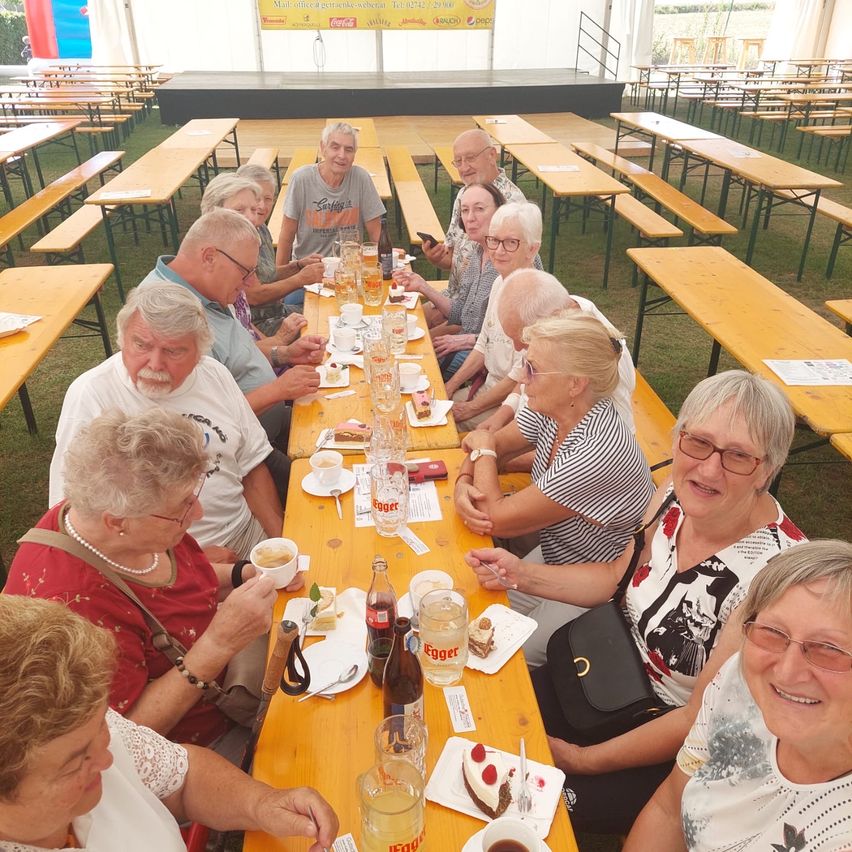 Eine Gruppe älterer Menschen sitzt an einem langen Tisch in einem Essbereich im Freien und genießt Getränke und Essen. Sie lächeln und scheinen eine gute Zeit zu haben.