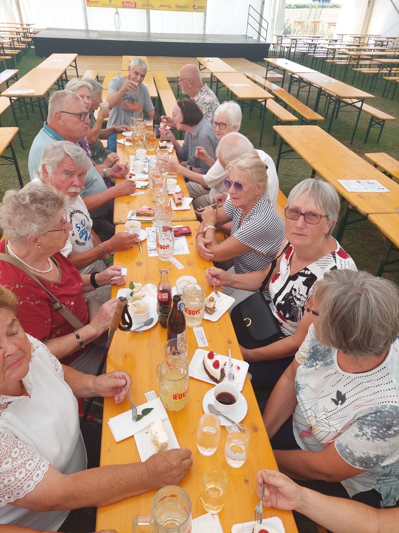 Eine Gruppe älterer Menschen sitzt an einem langen Tisch in einer Außenumgebung und genießt Essen und Getränke. Sie sind lässig gekleidet und einige tragen Brillen. Der Tisch ist mit Tellern, Gläsern und verschiedenen Speisen gedeckt. Im Hintergrund sind weitere Tische und Bänke zu sehen.