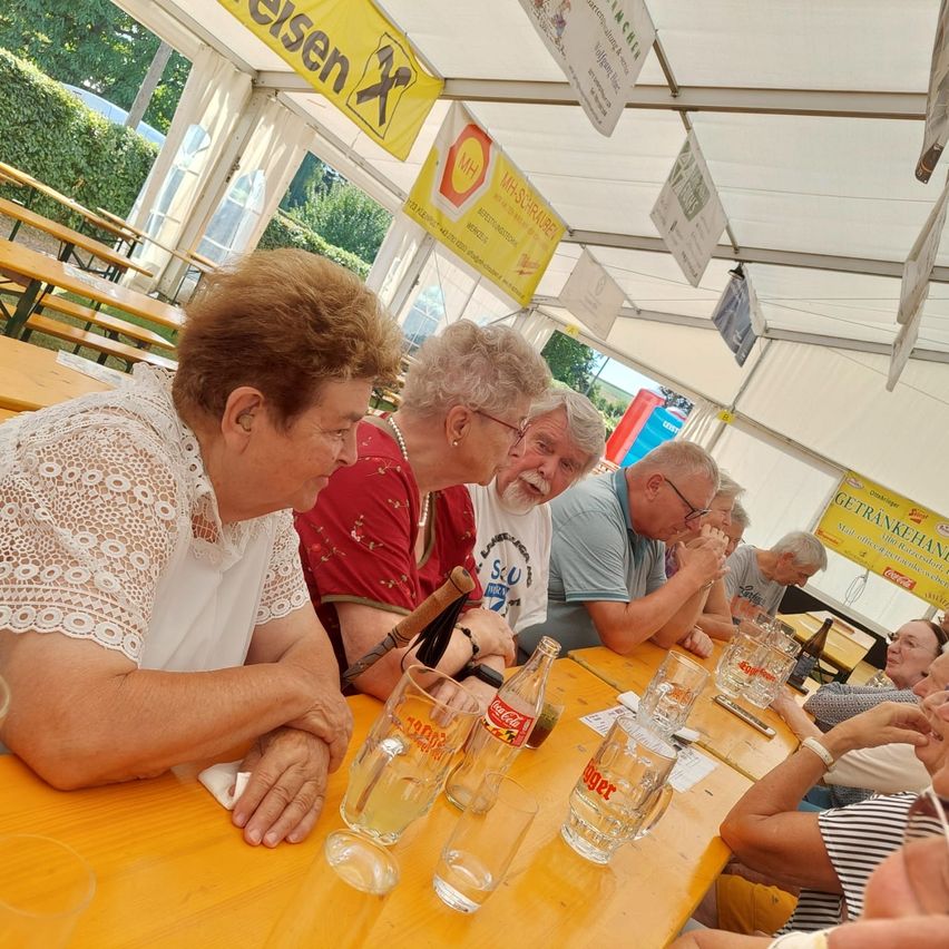 Eine Gruppe älterer Menschen sitzt unter einem Zelt an einem Tisch, einige mit Brille und einer hält einen Zigarren. Leere Gläser und eine Flasche sind auf dem Tisch.