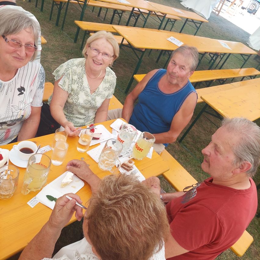 Eine Gruppe älterer Menschen sitzt an einem Tisch in einem Biergarten im Freien, lächelt und genießt ihre Getränke.