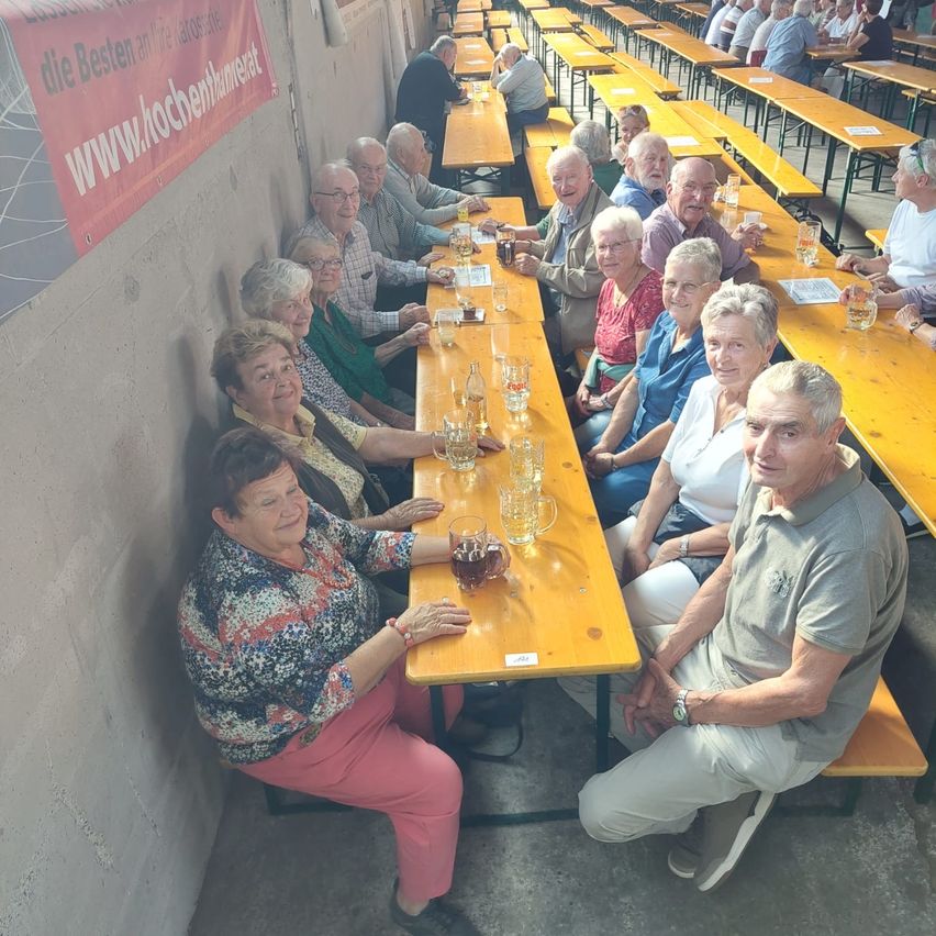 Eine Gruppe älterer Menschen sitzt an einem langen Holztisch in einem Restaurant, lächelt und posiert für ein Foto. Sie sind von anderen Tischen umgeben, an denen Menschen sitzen und stehen.