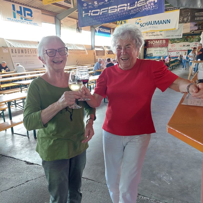 Zwei ältere Frauen lächeln und halten Weinglas in einer Biergarten. Sie stehen nebeneinander, wobei eine Frau ein grünes Shirt trägt und die andere ein rotes Shirt. Hinter ihnen sind Tische und Stühle, und in der Ferne sitzen und stehen Leute.