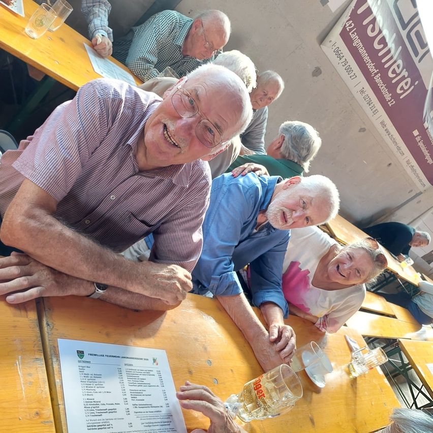 Eine Gruppe älterer Menschen sitzt an einem Tisch in einem Restaurant, lächelt und posiert für ein Foto. Der Tisch hat eine Speisekarte, Gläser und eine Flasche Bier. Im Hintergrund ist eine Wand mit einem Banner und eine verschwommene Ansicht anderer Menschen zu sehen.