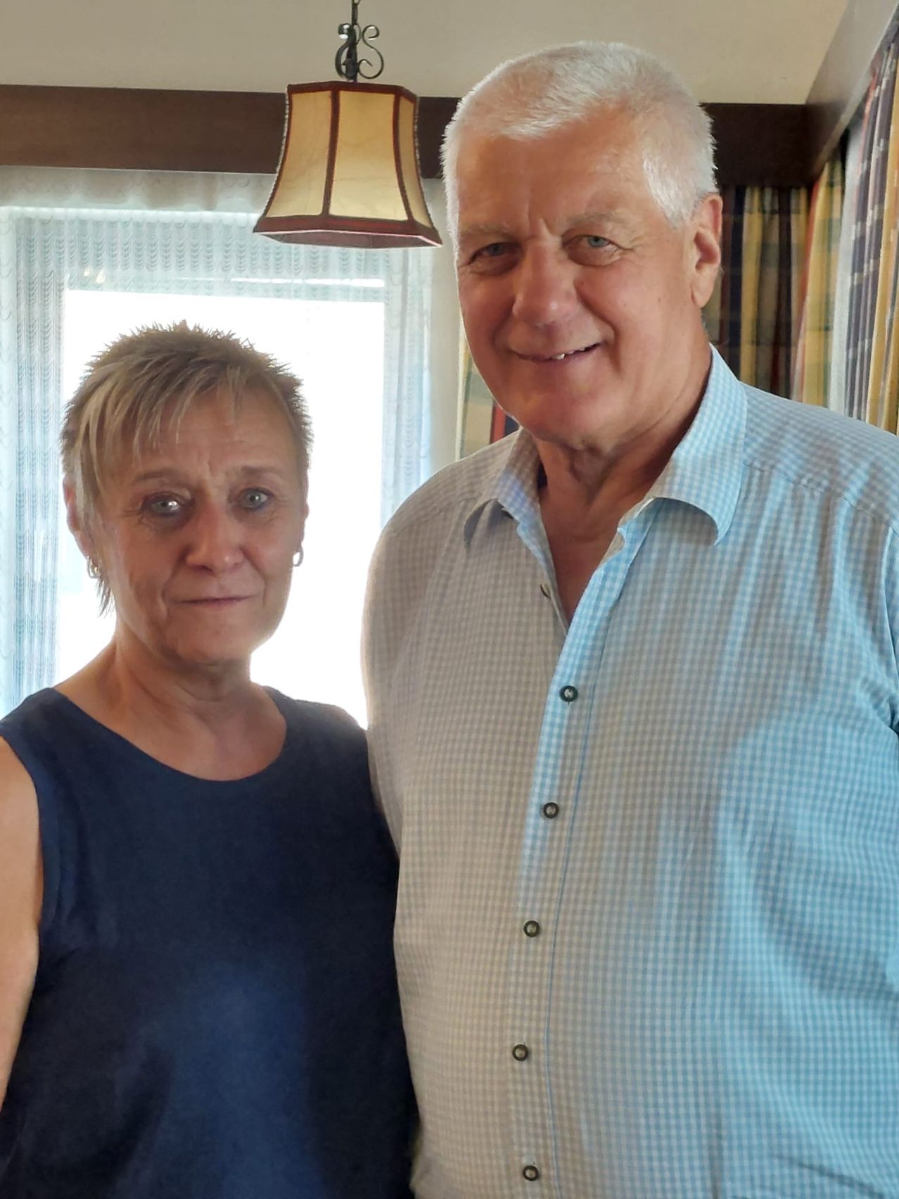 A man and woman pose for a picture in front of a window with white curtains. The man wears a blue checkered shirt, and the woman wears a blue sleeveless top.