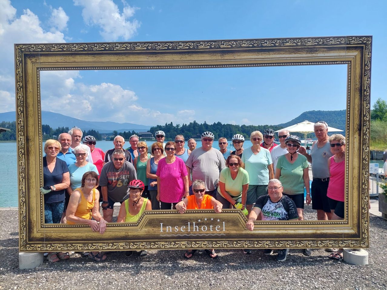 Eine Gruppe von Radfahrern posiert für ein Foto vor einem großen goldenen Rahmen vor einem See und Bergen.