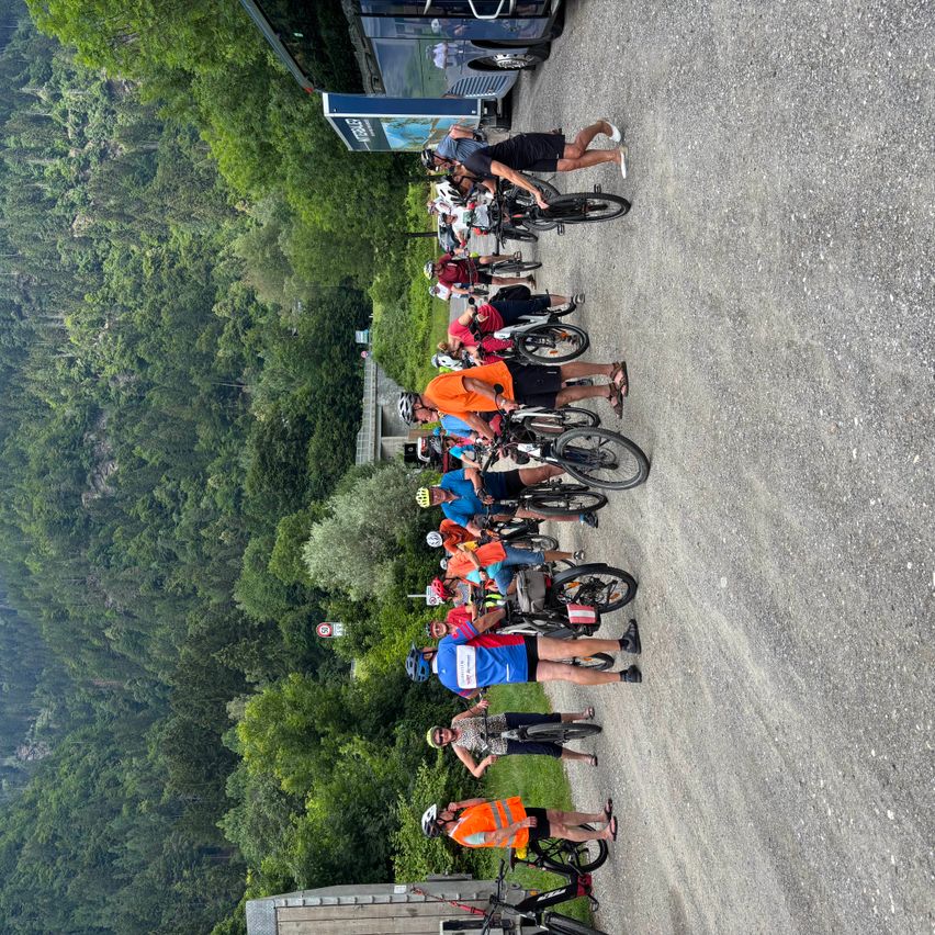Eine Gruppe von Radfahrern steht auf einem Schotterweg in einem bergigen Gebiet. Die Radfahrer sind in verschiedene Radsportbekleidung und Helme gekleidet. Bäume und Berge sind im Hintergrund.