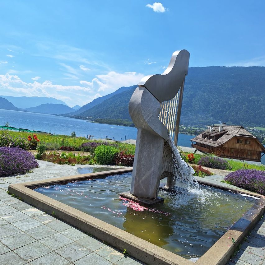 Ein Harfenförmiger Brunnen fließt in einen kleinen Pool, mit einem See und Bergen im Hintergrund.