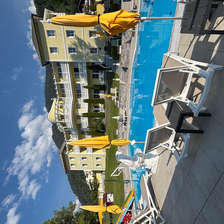Luftaufnahme eines Resorts mit Pool und gelben Sonnenschirmen, umgeben von grünem Rasen und Bäumen.