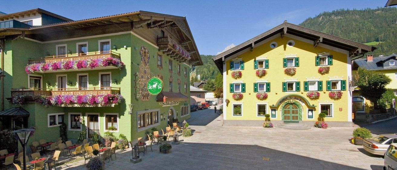 Zwei Gebäude mit Balkonen und Blumen, eines grün und eines gelb, mit einem Weg, Sitzgelegenheiten im Freien und einem Berg im Hintergrund.