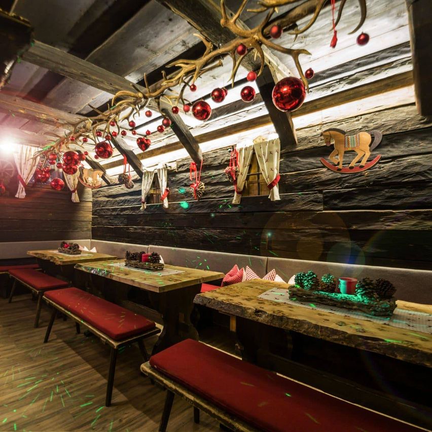 Ein Holzrestaurant mit Weihnachtsthema bietet rote Bänke und Tische, die mit Tannenzapfen und roten Kugeln geschmückt sind, und eine Wand, die mit Weihnachtskugeln und einem Schaukelpferd verziert ist.