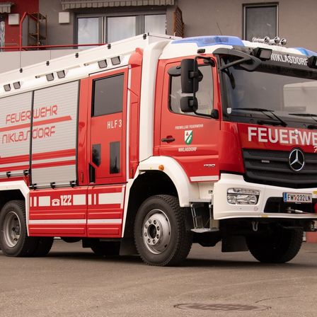Ein roter und weißer Mercedes-Benz Feuerwehrwagen steht vor einem Gebäude mit Balkon. Der Wagen ist als HLF 3 gekennzeichnet.