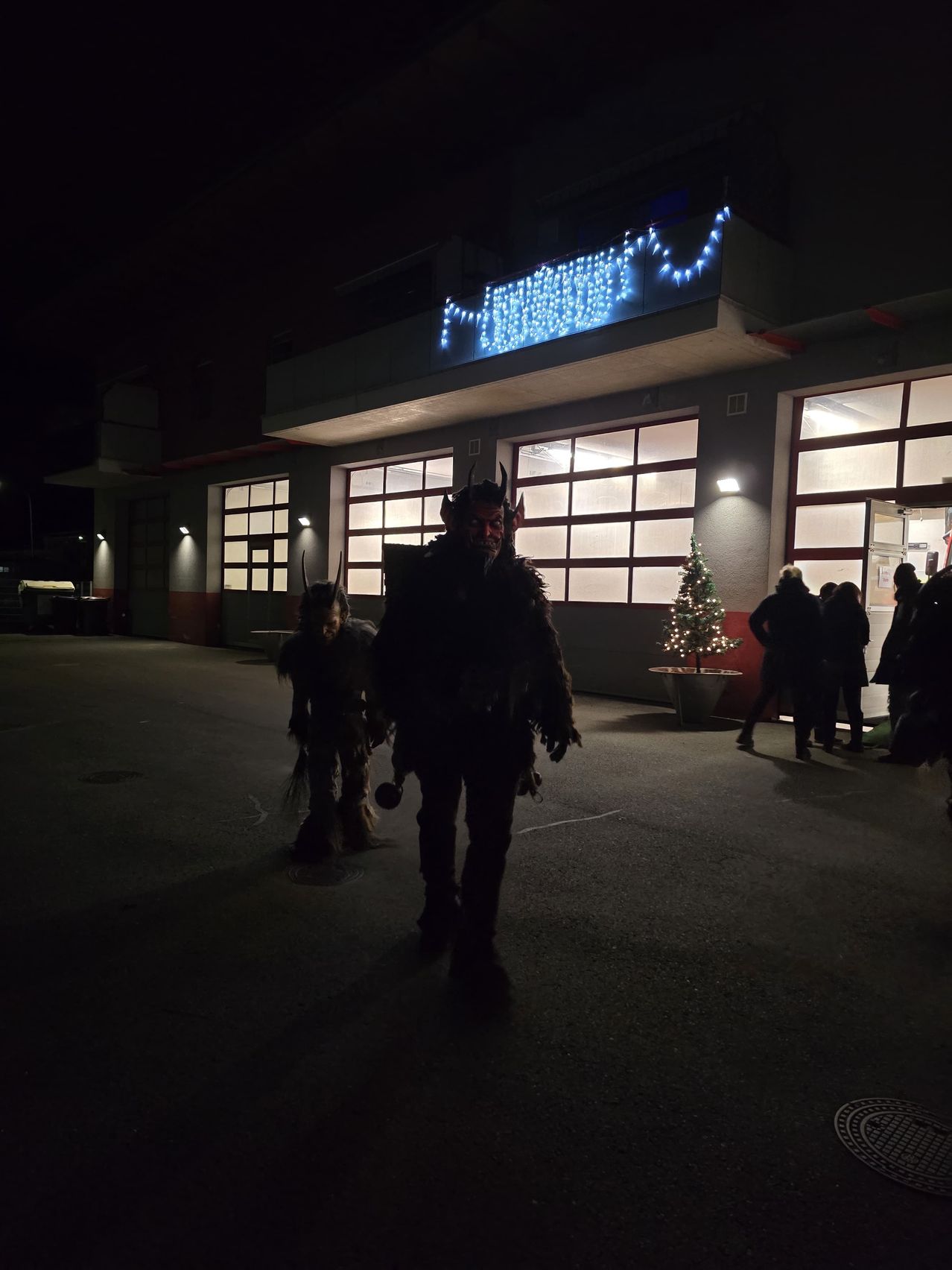 Zwei Personen in Kostümen gehen vor einem Gebäude mit einem Weihnachtsbaum und Lichtern. Einer trägt ein Teufelskostüm, der andere ist kleiner.