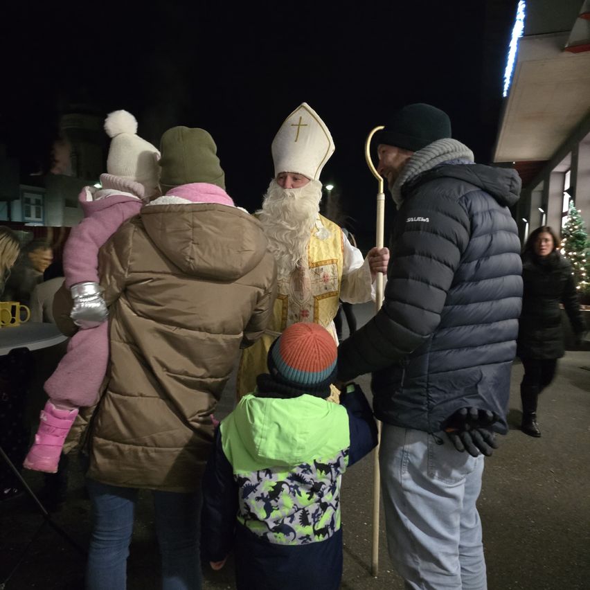 Ein Priester in festlicher Kleidung steht mit einem Stab und begrüßt eine Familie und ein Kind an einem geschäftigen Ort im Freien bei Nacht.
