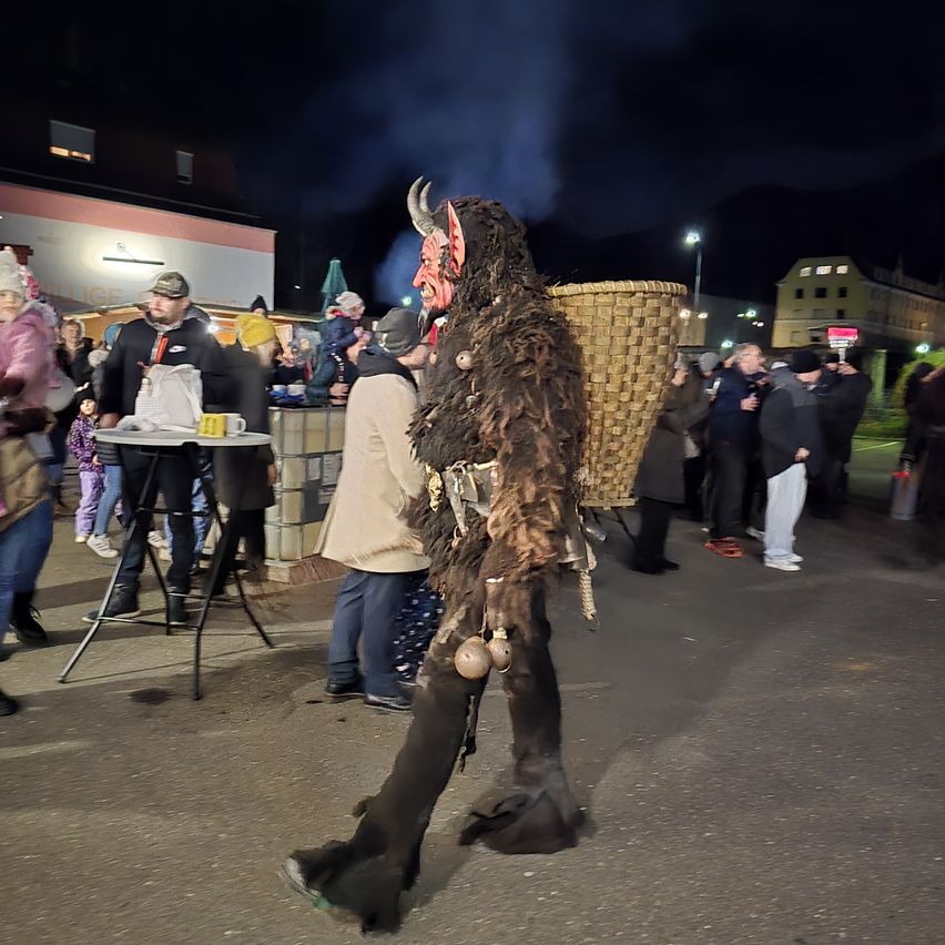 Eine Person in einem gruseligen Kostüm geht durch eine Menschenmenge auf einer Nachtstraße. Im Hintergrund steht ein Tisch mit Tassen und Tellern.