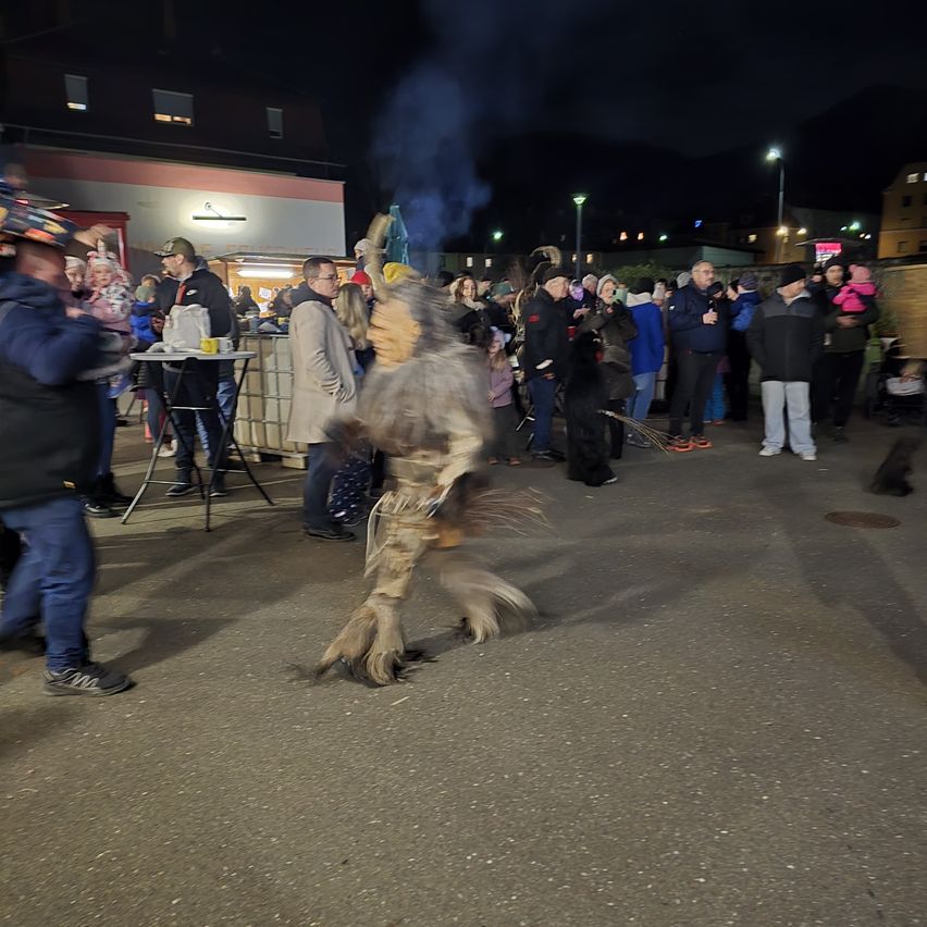 In der Nacht tanzt eine Person in einem pelzigen Kostüm vor einer Menschenmenge, wobei Rauch hinter ihr aufsteigt. Leute in Jacken stehen herum und schauen zu. Ein Tisch mit Tassen und Flaschen steht in der Nähe.