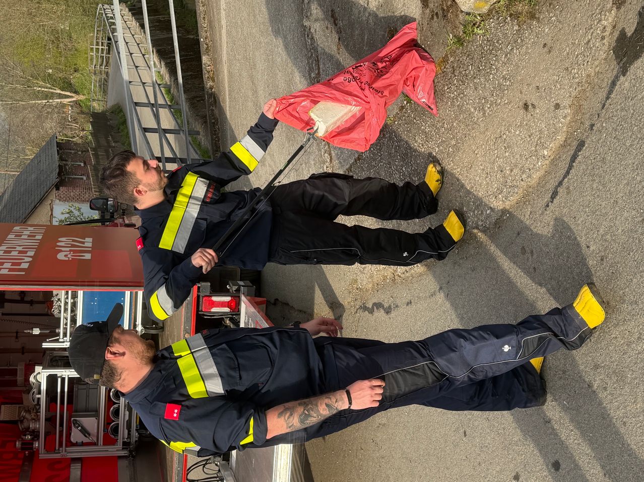 Zwei Feuerwehrleute in Uniform sind auf einer Straße, einer hält einen roten Beutel. Sie sind in der Nähe eines Feuerwehrwagens mit einer roten Tür und einer Brücke im Hintergrund.