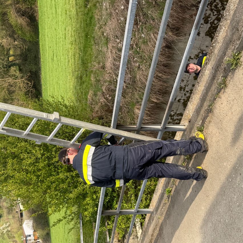 Zwei Männer in Schutzausrüstung befinden sich in der Nähe eines Baches. Einer steht am Rand, der andere ist teilweise eingetaucht. Eine Leiter lehnt an einem Metallzaun.