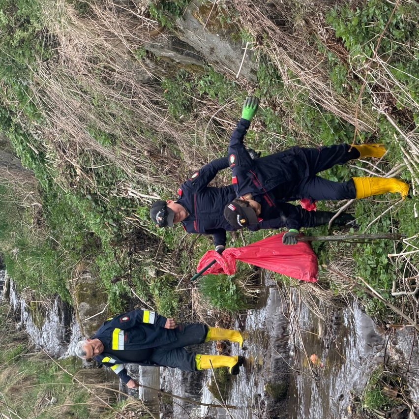 Drei Personen in Schutzkleidung befinden sich in der Nähe eines Baches und sammeln Müll. Eine Person ist am Ufer, eine andere liegt mit einer roten Tasche und die dritte steht.