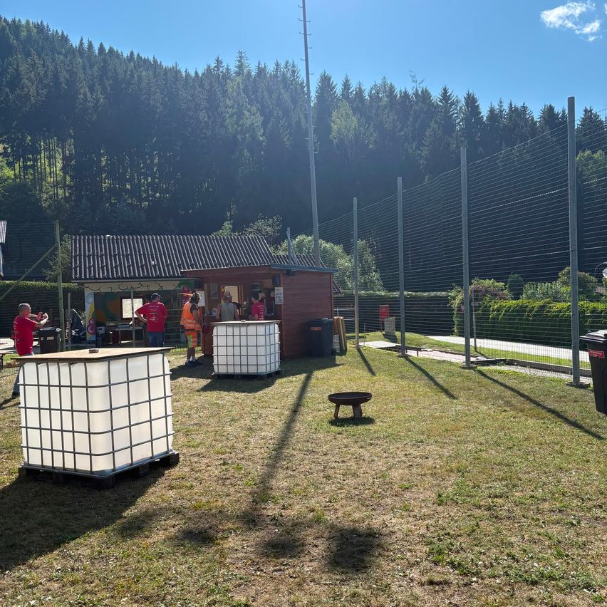 Eine Gruppe von Menschen steht in der Nähe eines kleinen Gebäudes mit einem Zaun, und ein Mülleimer ist in der Nähe. Eine Person steht links, und einige Leute stehen vor dem Gebäude. Einige Lagerboxen sind auf dem Boden platziert. Ein Mülleimer ist rechts platziert.