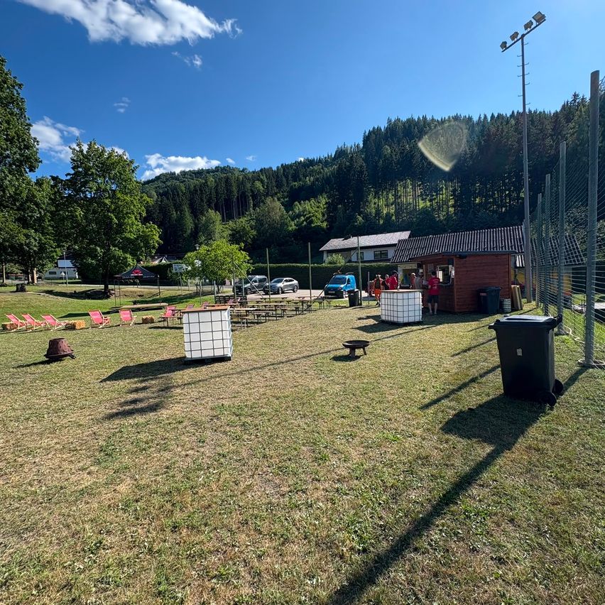Ein Außenbereich mit einem Grasfeld, einem kleinen Gebäude und mehreren Personen. Es gibt Tische, Stühle und Lagerboxen verstreut. In der Ferne gibt es Bäume und einen Berg.