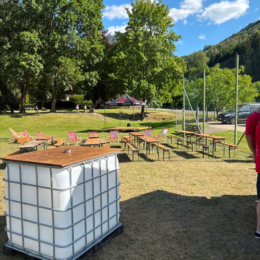 Ein Außenbereich mit Picknicktischen, Bänken und einem Baldachin. Ein großer weißer Lagercontainer steht im Vordergrund. Bäume und ein Berg sind im Hintergrund.