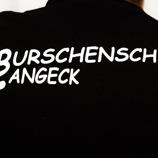 Ein schwarzes Shirt zeigt weißen Text, der 'BURSCHENSCHAFT ANGECK' liest. Das Design ist fett und zentriert auf dem Rücken.
