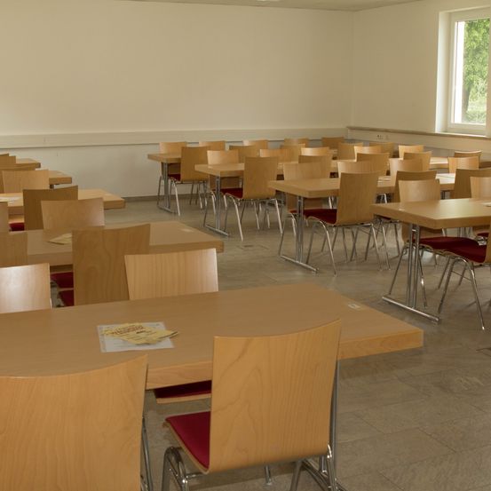 Ein leerer Klassenraum mit mehreren Holztischen und Stühlen. Ein Tisch im Vordergrund hat ein Buch darauf. Der Raum hat eine weiße Wand und ein Fenster mit Blick auf Bäume.