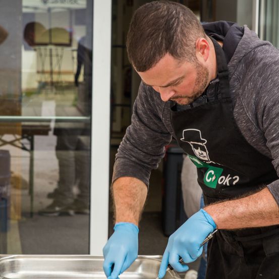 Ein Mann in einem Küchenschürze bereitet Essen zu und trägt blaue Handschuhe. Er steht vor einer Edelstahlschale. Hinter ihm befindet sich eine Glastür mit einer verschwommenen Reflexion einer Person.