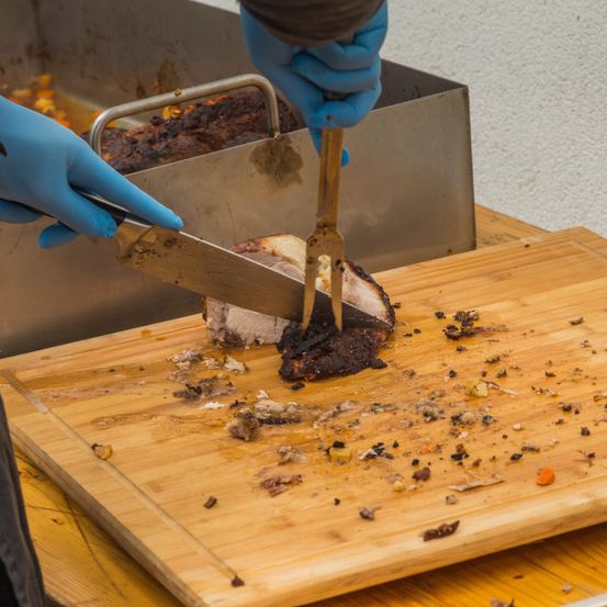 Eine Person mit blauen Handschuhen schneidet ein Stück Fleisch auf einem hölzernen Schneidebrett mit Messer und Gabel. Das Fleisch liegt auf einem Edelstahltablett mit Griff.