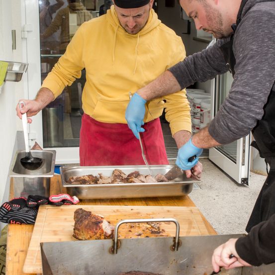 Zwei Männer in einer Küche, einer trägt eine gelbe Kapuzenjacke und eine rote Schürze, der andere ein graues Shirt, beide benutzen blaue Handschuhe, um Essen zuzubereiten.