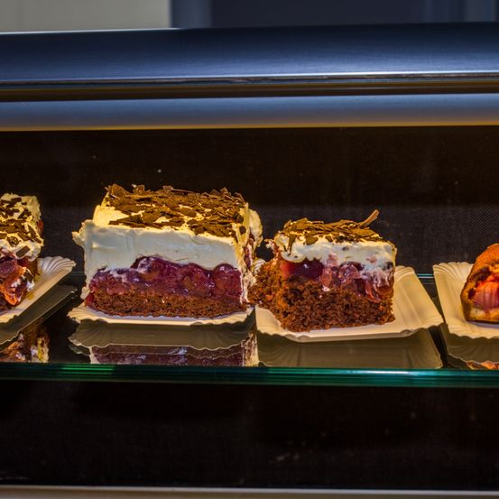 Eine Vitrine mit vier Desserts. Drei sind geschichtete Kuchen mit weißer Glasur und dunklen Schokoladenraspeln. Das vierte Dessert ist eine Backware mit roter Füllung.