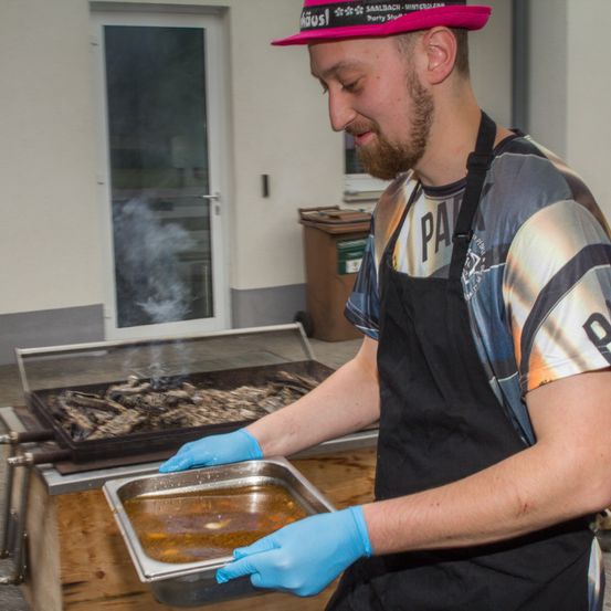 Ein Mann mit einem rosa Hut und blauen Handschuhen kocht draußen auf einem Grill Essen.
