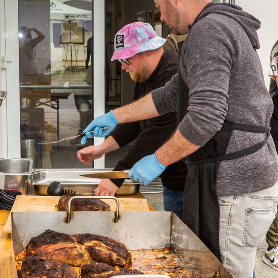 Zwei Männer schneiden Fleisch auf einem Schneidebrett. Einer trägt einen rosa Hut und der andere einen grauen Kapuzenpullover. Sie stehen vor einem Tresen mit einer Edelstahlschale.