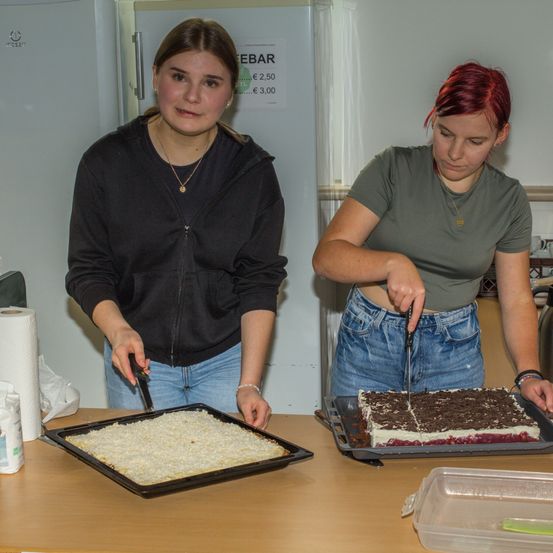 Zwei Frauen in einer Küche bereiten Essen zu. Eine hat einen Tablett mit Kuchenkrümeln, und die andere schneidet einen Kuchen auf einem anderen Tablett. Im Hintergrund steht ein Kühlschrank mit aufgelisteten Preisen.