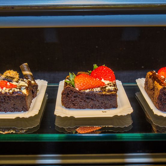 Drei Schokoladen-Brownies sind ausgestellt. Einer hat Erdbeeren und Schlagsahne, ein anderer hat eine Schokoladenglasur und der dritte hat Nüsse obenauf.