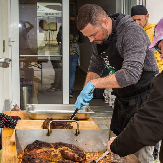 Ein Mann in einer grauen Kapuzenjacke und blauen Handschuhen bereitet Fleisch in einer Schale zu. Eine andere Person in einer schwarzen Jacke steht neben ihm und beide konzentrieren sich auf die Aufgabe. Im Hintergrund spiegelt eine Glastür eine Person und ein Zelt.