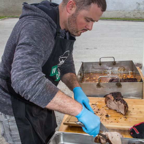Ein Mann in einer grauen Kapuzenjacke und Schürze schneidet Fleisch auf einem Schneidebrett. Er trägt blaue Handschuhe und steht neben einem Tisch mit einem Tablett mit Fleisch. Im Hintergrund sind eine Wand und eine Betonstraße zu sehen.
