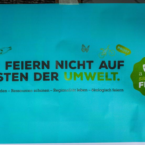 Ein Poster mit blauem Hintergrund zeigt eine Botschaft zum Umweltschutz. Es steht 'Feiern nicht auf der Umwelt.' Symbole einer Schmetterling, eines Fahrrads und einer Flasche sind vorhanden. Der Text ermutigt auch zur Einsparung von Ressourcen, regionalem Leben und umweltfreundlichen Feiern.