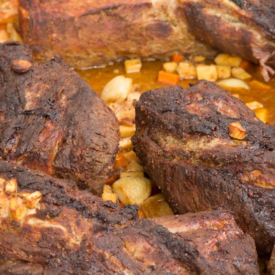 Eine Nahaufnahme von verkohlten Fleischstücken, möglicherweise Rippen, mit karamellisierten Gemüsestücken im Hintergrund. Das Fleisch hat eine knusprige, rauchige Textur.