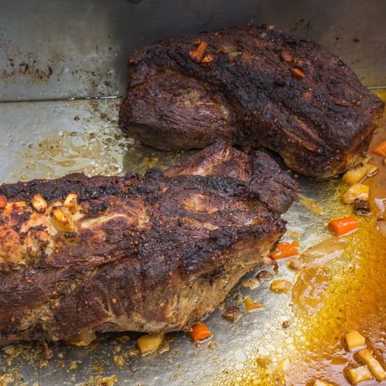 Zwei geräucherte Fleischstücke, wahrscheinlich Schweinefleisch, liegen auf einem Metalltablett mit Säften und Gemüse. Ein Stück hat eine verkohlte Oberfläche und eine Garnitur.