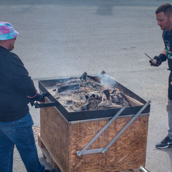 Zwei Männer auf einem Parkplatz steuern eine große Holzkiste mit offenem Feuer darin, einer mit einer Schaufel und der andere mit einem Stock.
