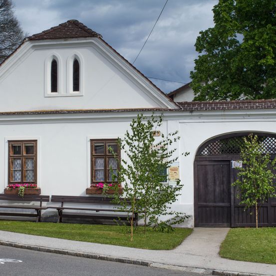 Ein weißes Haus mit einem braunen Dach, zwei Bogenfenstern und zwei Fensterbänken mit rosa Blumen. Ein Holztor führt zum Haus.