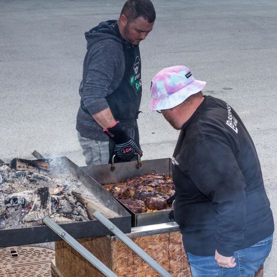 Zwei Männer grillen Fleisch im Freien. Einer trägt eine Tie-Dye-Kappe und Handschuhe, der andere eine Kapuzenjacke mit grünem Logo. Das Fleisch liegt auf einem Grill mit offenem Feuer.