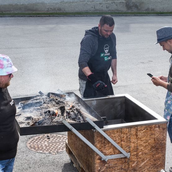 Drei Männer kümmern sich um einen großen Outdoor-Grill mit brennenden Kohlen. Einer benutzt ein Telefon, ein anderer hält eine Pfanne und der dritte steht in der Nähe eines Mannlochdeckels.