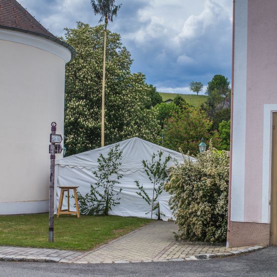 Ein weißes Zelt steht in einem Hof mit einem Holzhocker daneben. Ein Gebäude mit einem braunen Dach befindet sich links, und ein Busch mit Blumen rechts. Bäume und ein bewölkter Himmel sind im Hintergrund.